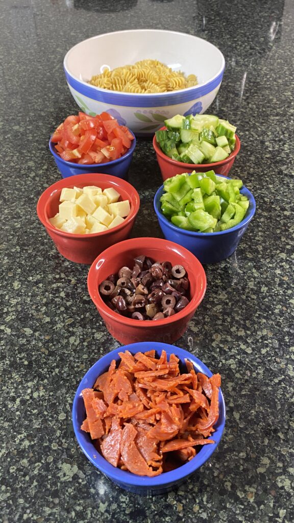 Perspective shot 2 on pepperoni, cheese, green pepper, tomatoes, cucumber, and Kalamata olives in serving bowls.  